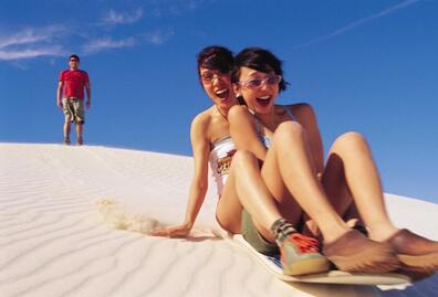 Two backpackers sandboarding down the white sand dunes of Lancelin, Western Australia, enjoying a thrilling outdoor adventure on a sunny day.